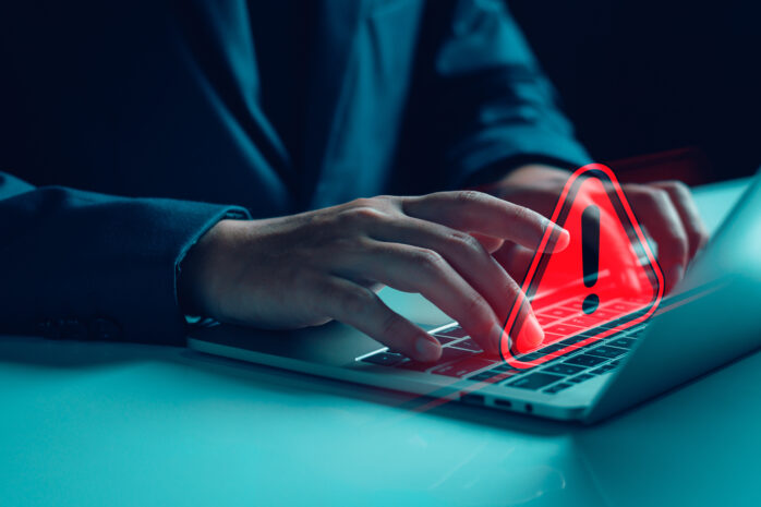 A close-up of a person's hands typing on a laptop with a red warning symbol overlay, representing a cybersecurity threat.