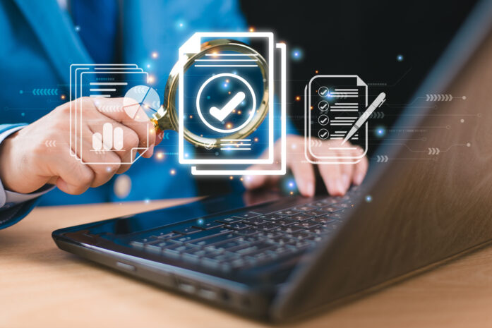 A professional using a magnifying glass over digital audit icons displayed on a laptop screen, symbolizing IT audits, data security, and compliance.